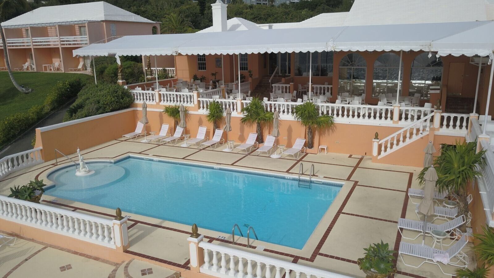 A pool area with lounge chairs and umbrellas, surrounded by a white fence and a building with balconies.