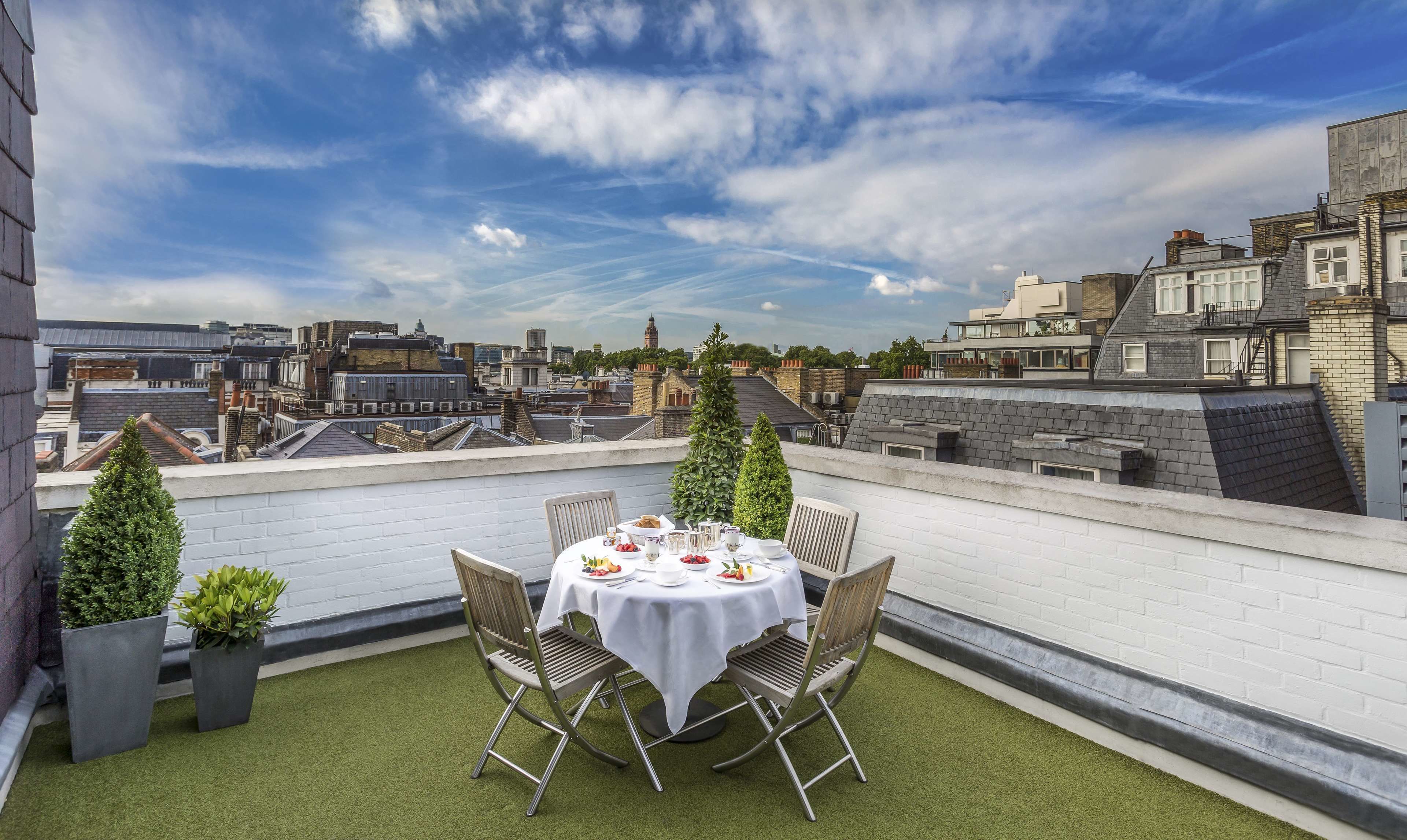 A rooftop terrace with a table set for two, surrounded by potted plants, overlooking a cityscape with historic buildings.