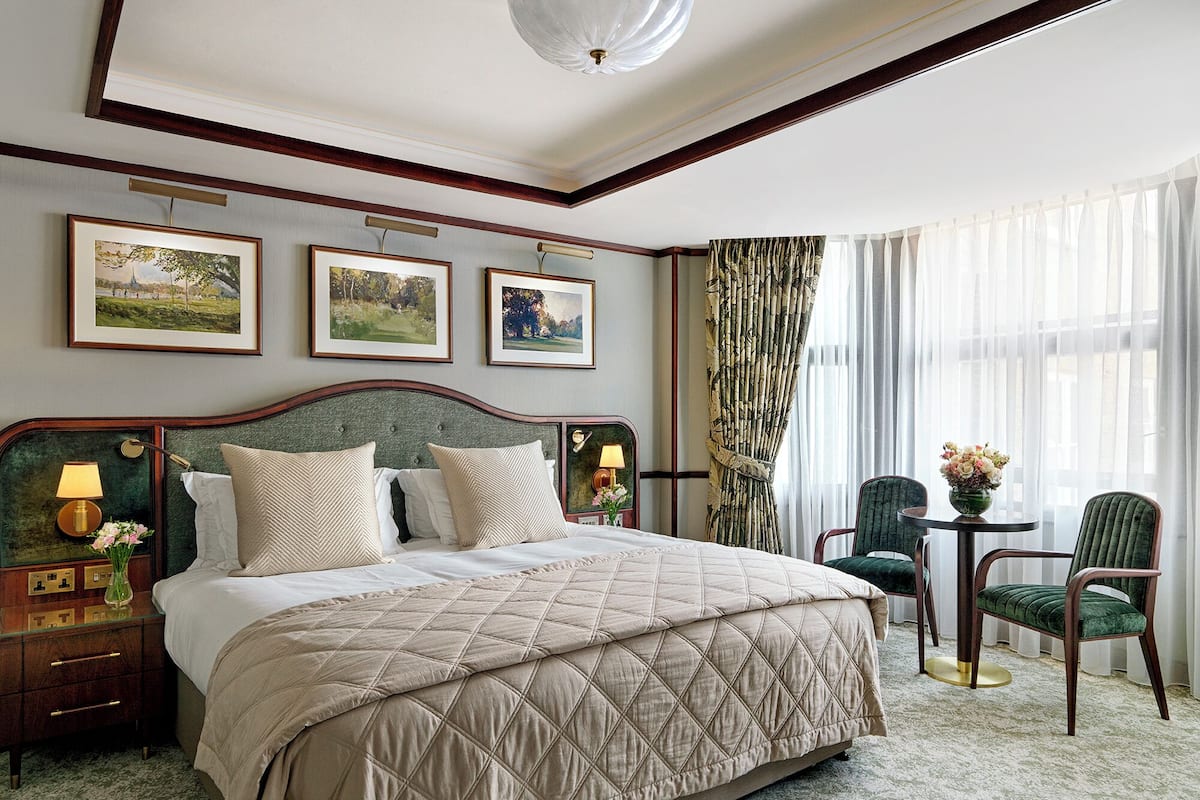 A hotel room with a large bed, a nightstand with a lamp, a chair, and a table with a vase of flowers.