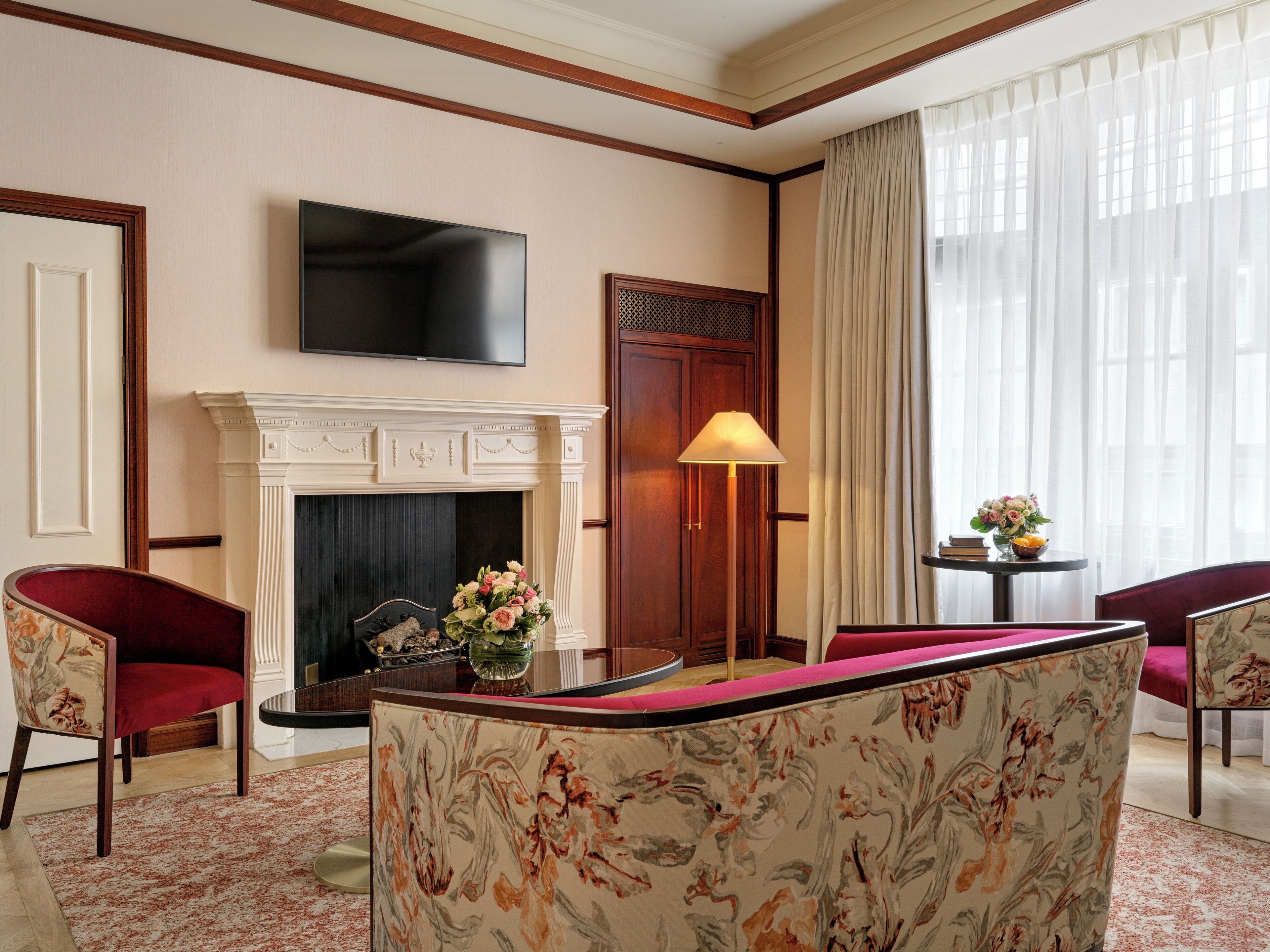 A hotel room with a fireplace, a television mounted above it, a desk with a vase of flowers, and two red upholstered chairs.