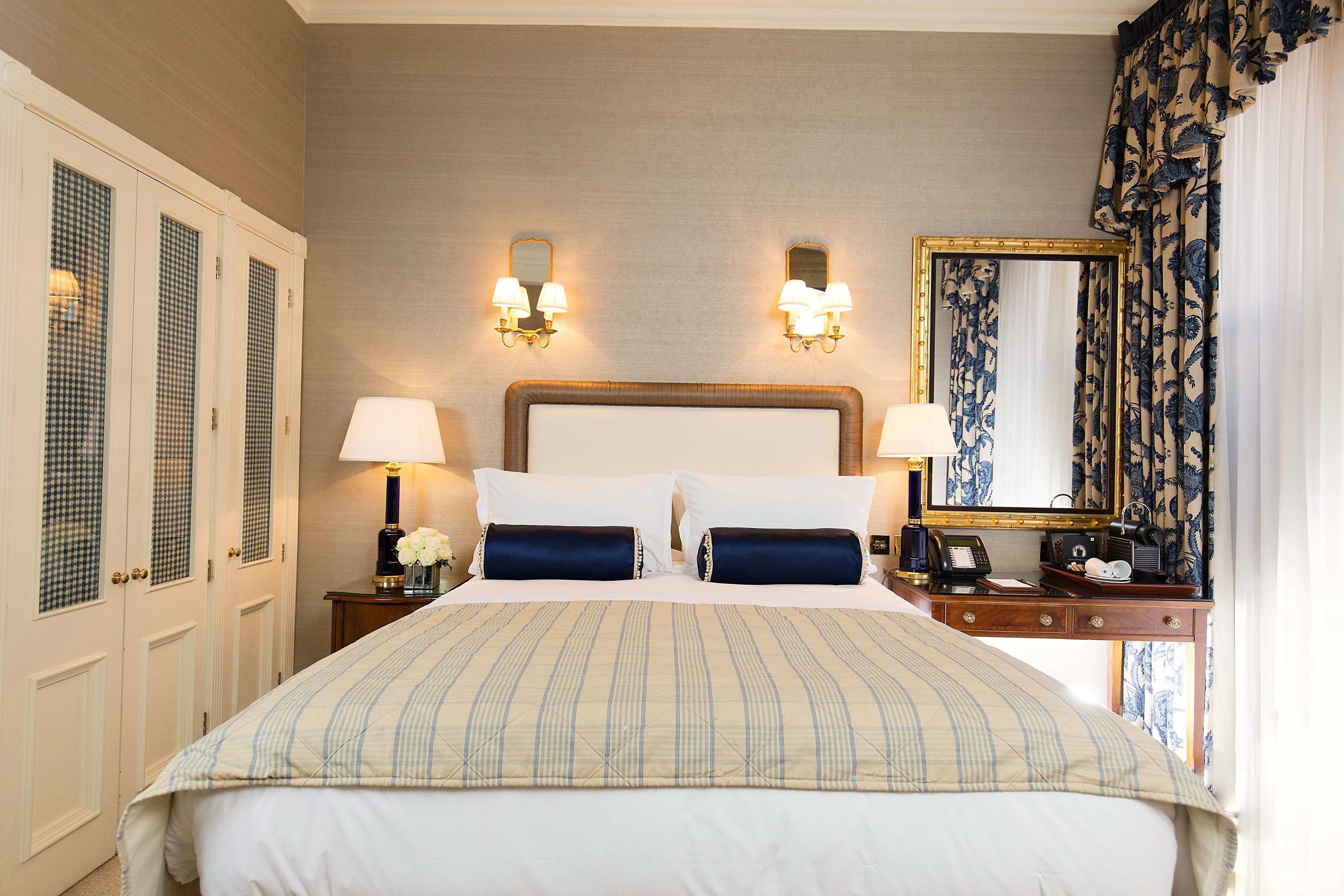 Habitación de hotel con una cama grande, dos lámparas de noche, una mesita con teléfono, un espejo y una ventana con cortinas.