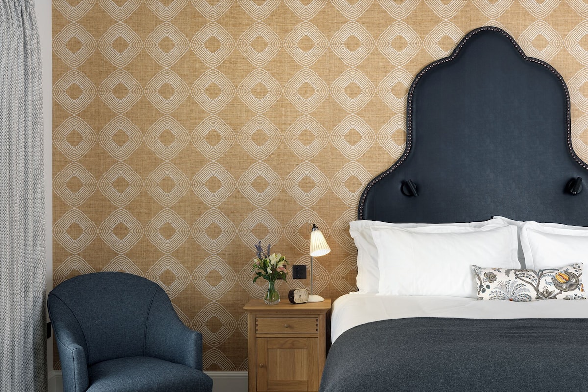 A bedroom with a patterned wallpaper, a dark headboard, a wooden nightstand with a lamp, a blue armchair, and a bed with white bedding and a decorative pillow.