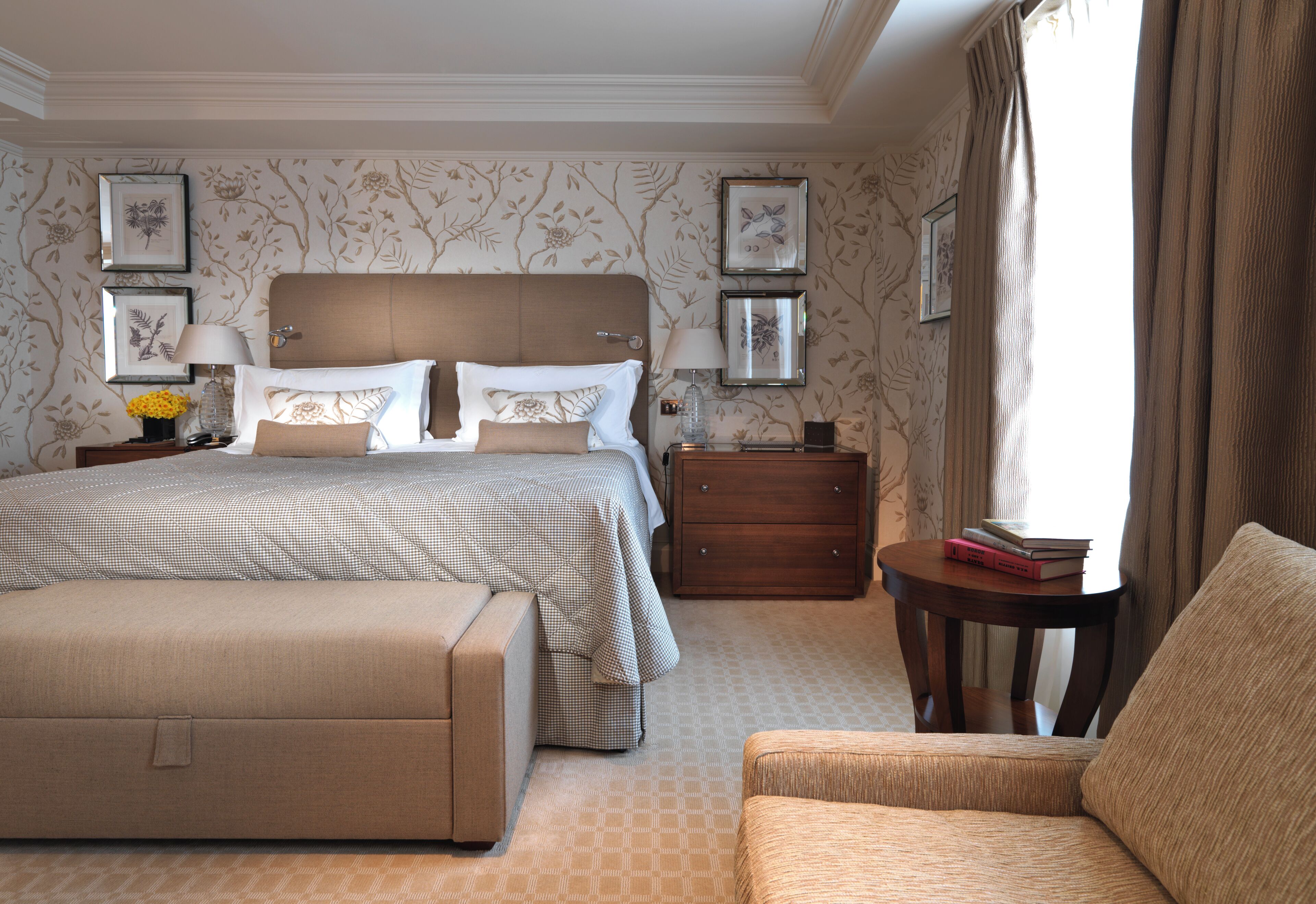 Quarto de hotel com uma cama grande, uma mesa de cabeceira, uma cadeira e uma pequena mesa com livros.
