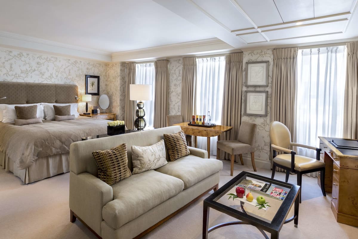 A hotel room with a large bed, a sofa, a desk, and a dining table. There are curtains, framed pictures on the walls, and a carpeted floor.