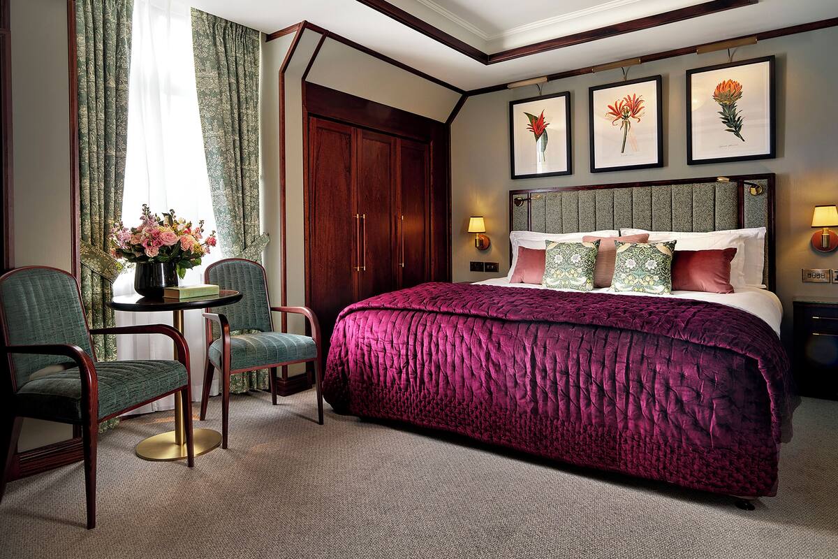 A hotel room with a large bed, two chairs, a table with a vase of flowers, and framed pictures on the wall.