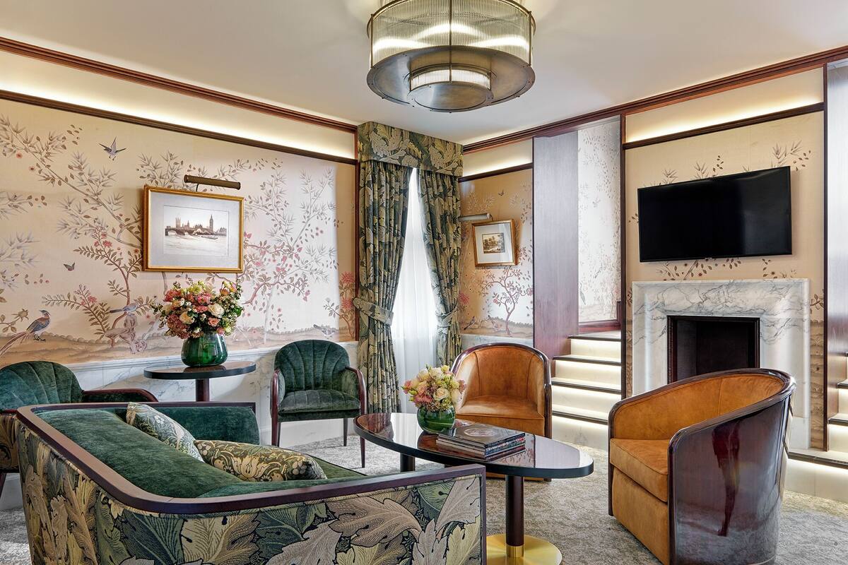 A spacious living room with a green sofa, a brown armchair, and a glass coffee table. There is a fireplace with a marble surround, and a flat-screen TV mounted above it.