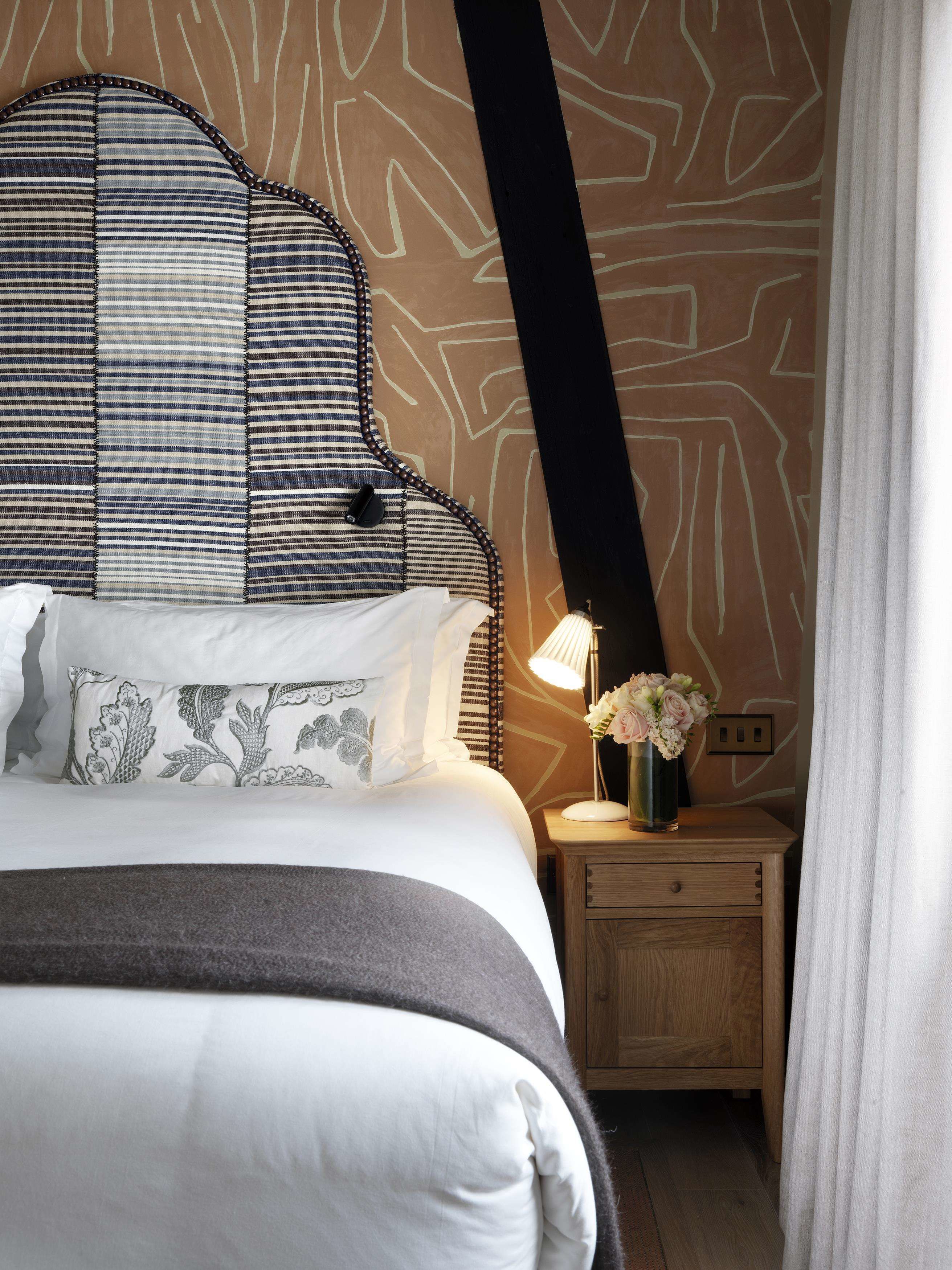 A neatly made bed with striped pillows, a bedside lamp, and a vase with flowers on a wooden nightstand.