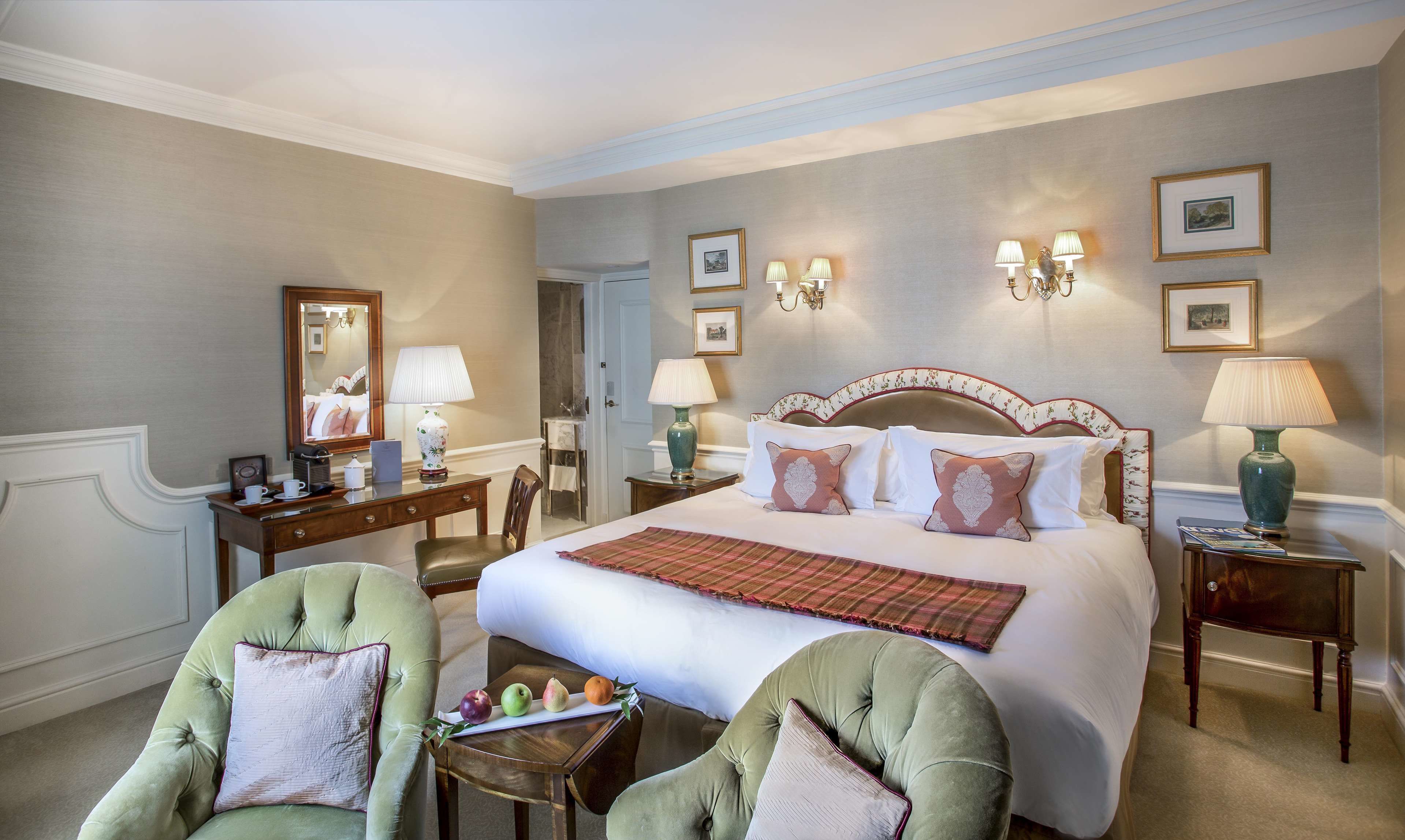 A hotel room with a large bed, two armchairs, a desk with a lamp, and a mirror.