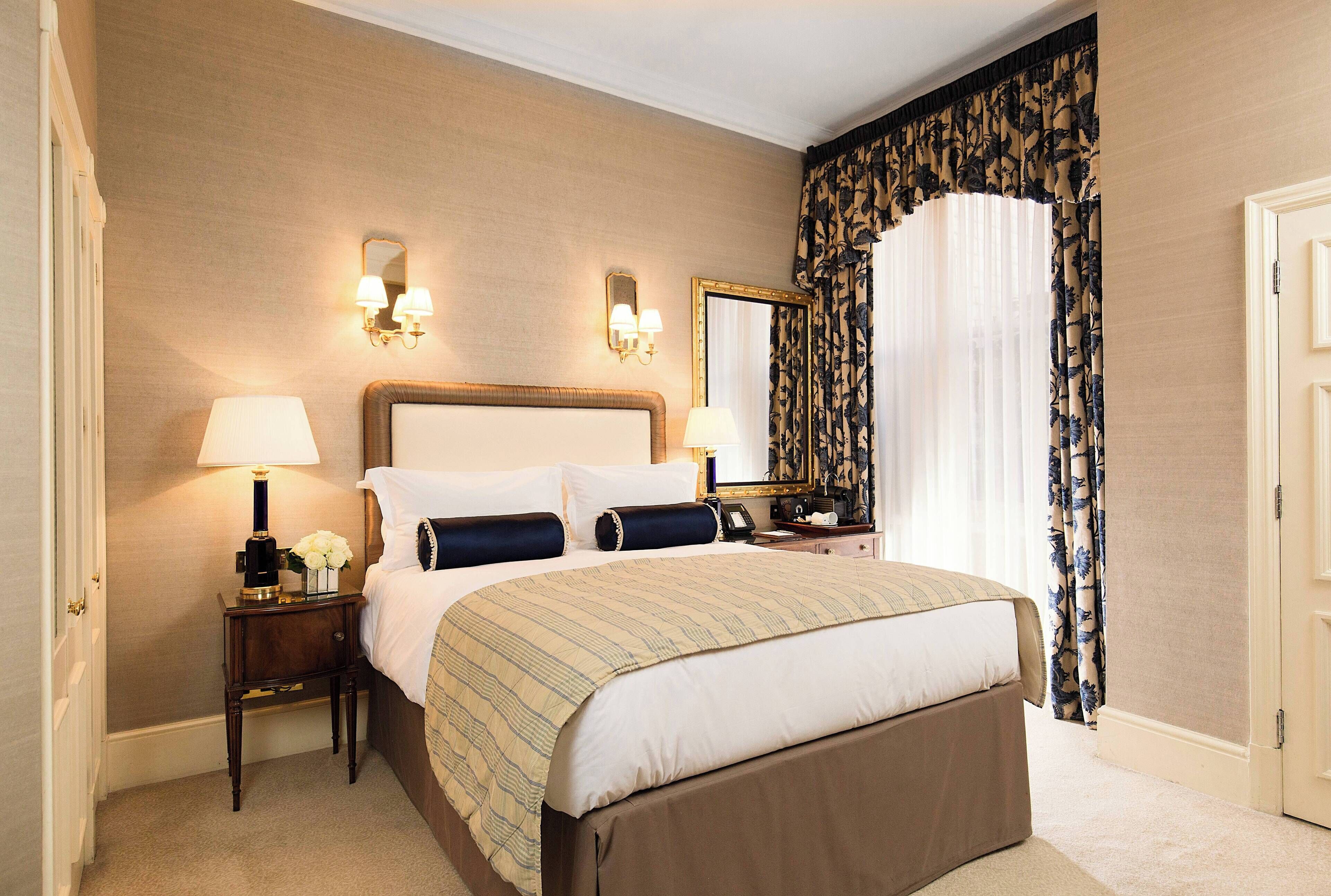 Une chambre d’hôtel avec un grand lit, une table de chevet avec une lampe, un miroir et une fenêtre avec des rideaux.