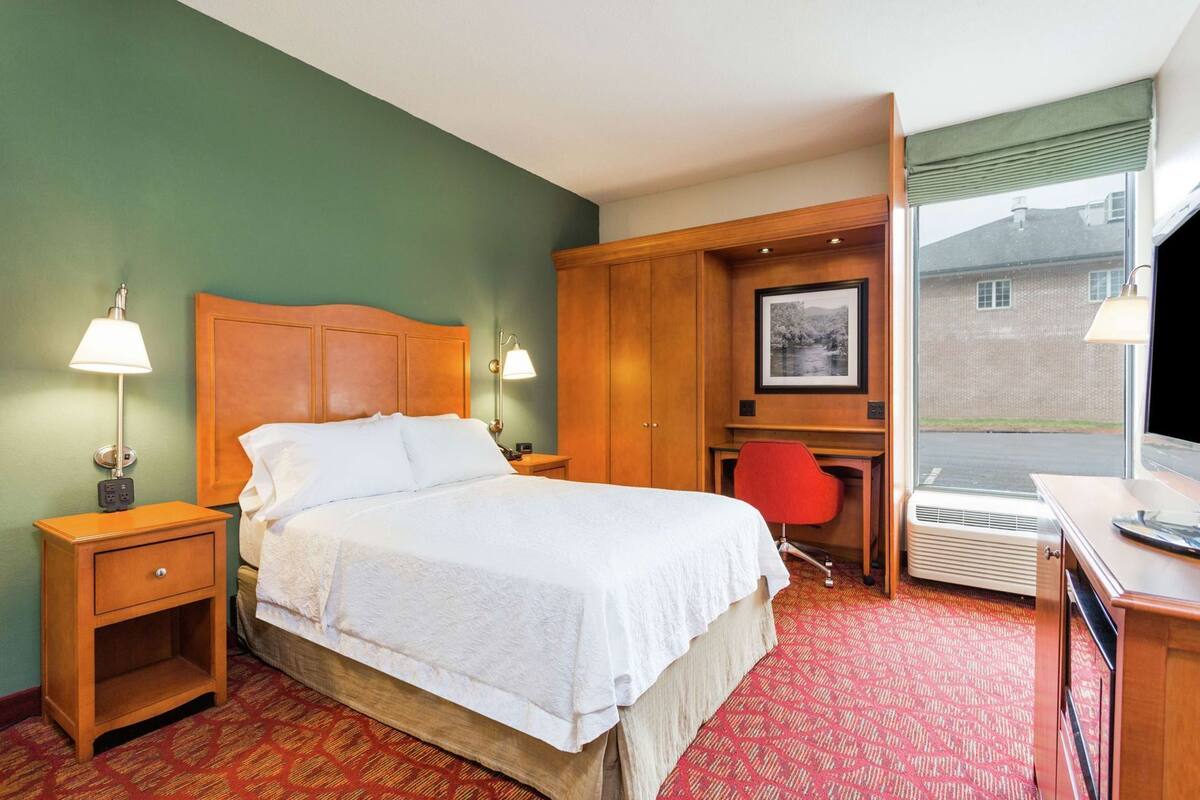 A hotel room with a bed, bedside tables, a desk, a chair, a window, and a picture on the wall.