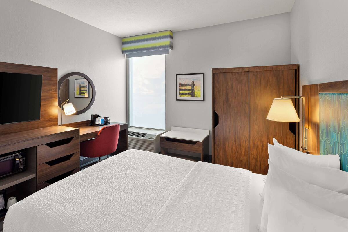 A hotel room with a bed, a desk, a chair, a TV, a mirror, a window, and a door.