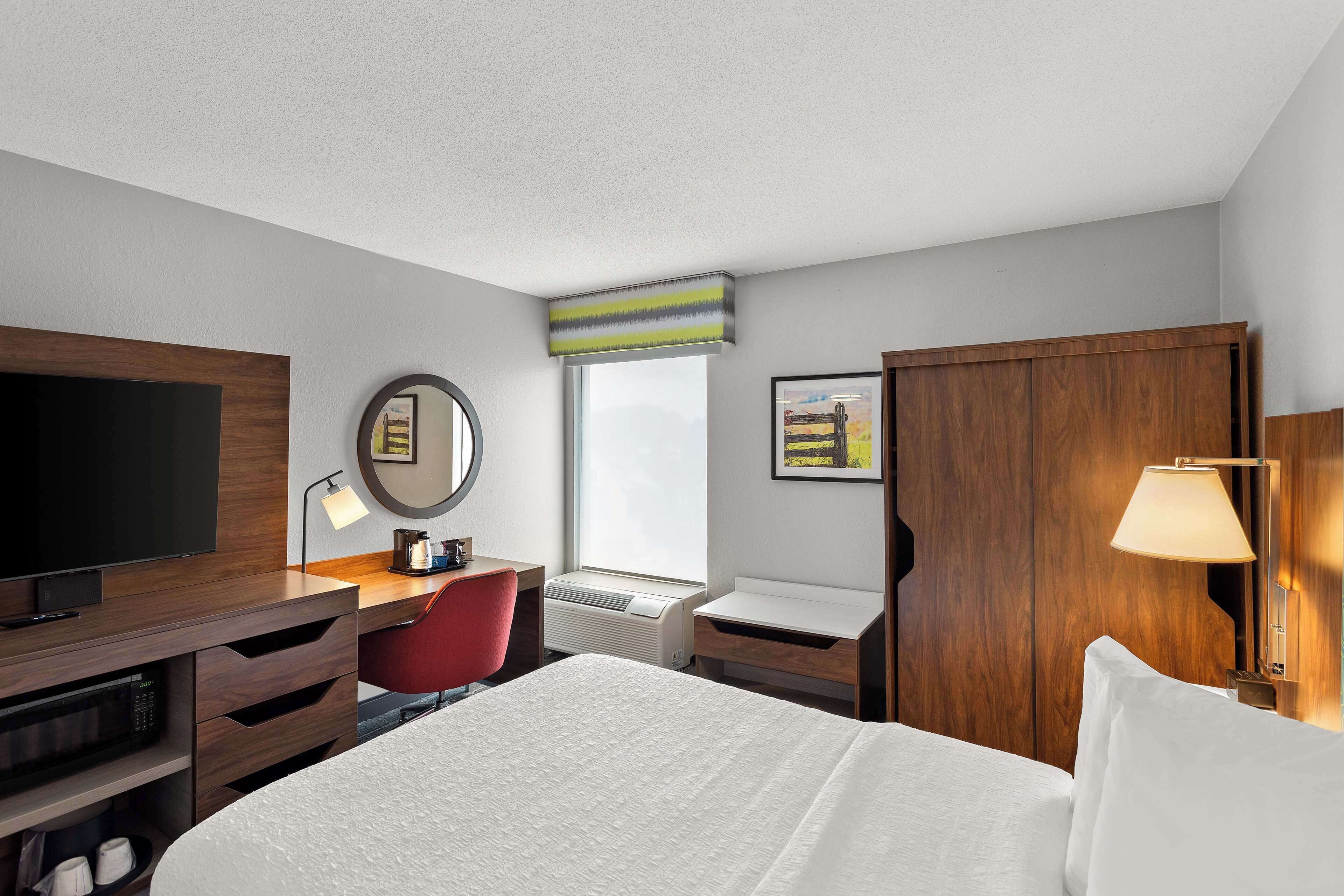 Une chambre d’hôtel avec un lit, un bureau avec une télévision, une chaise rouge, une armoire en bois et une fenêtre donnant sur un paysage agréable.