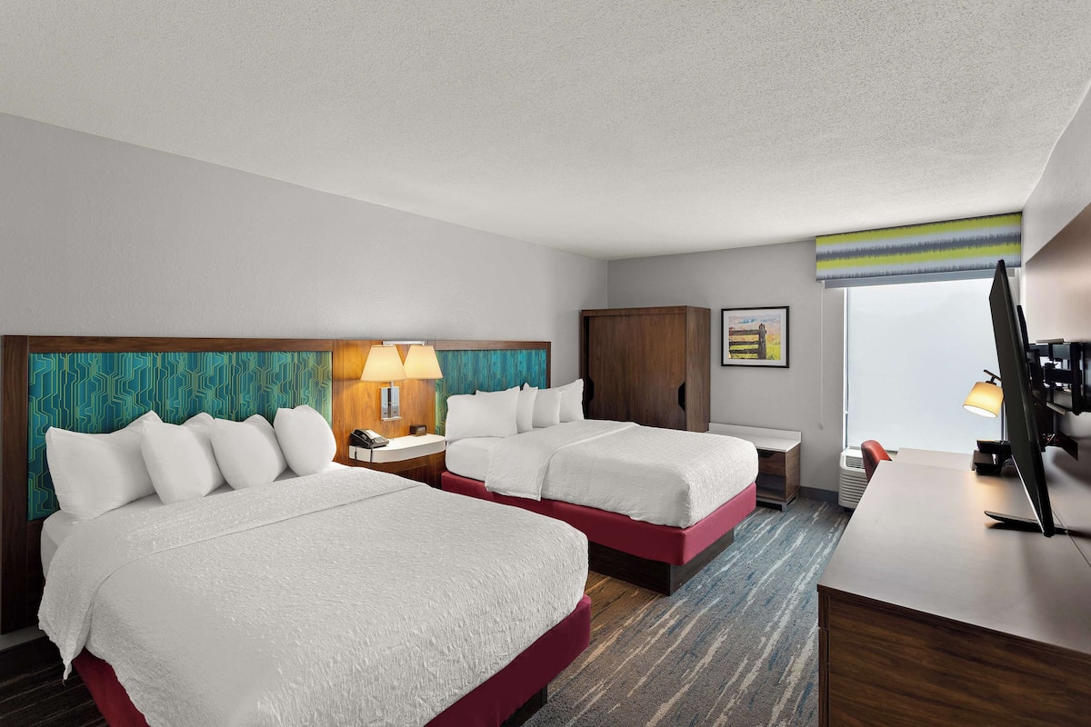A hotel room with two beds, a desk, a chair, a TV, and a window with a view.