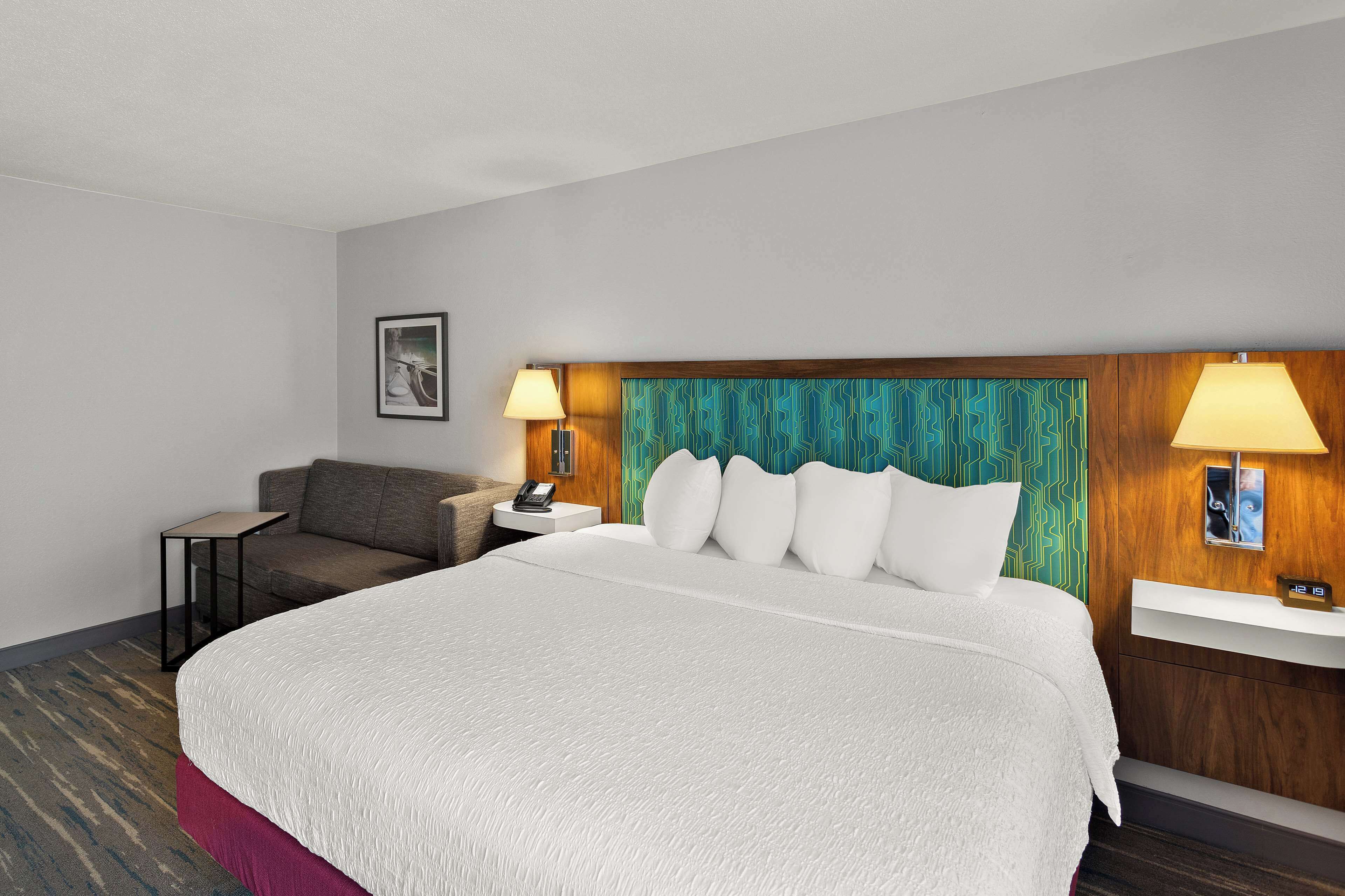 A hotel room with a large bed, a bedside table with a lamp, a small table, a sofa, and a wall-mounted shelf.