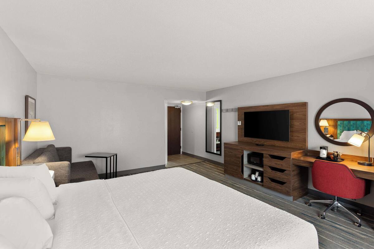 A hotel room with a large bed, a desk with a chair, a TV, a mirror, and a small table with a lamp.