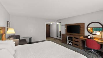Camera d'albergo con un letto grande, una scrivania con una sedia, una TV, uno specchio e un tavolino con una lampada.