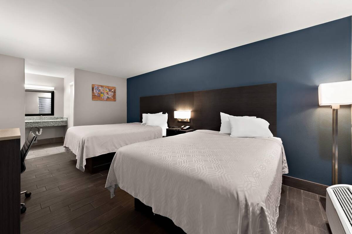 A hotel room with two beds, a desk, a chair, a nightstand, a lamp, and a wall-mounted artwork.