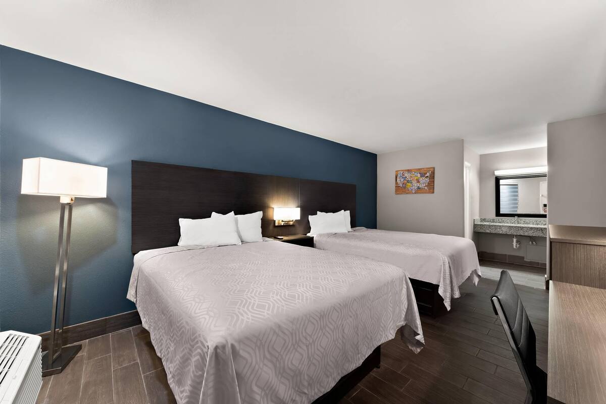 A hotel room with two beds, a nightstand with a lamp, a bathroom with a mirror, and a small seating area.