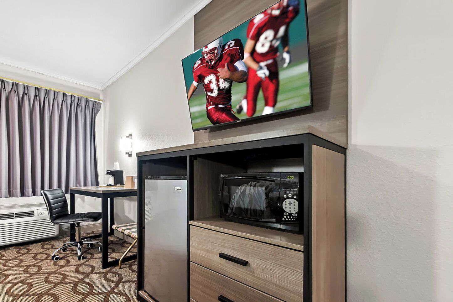 Habitación de hotel con televisión de pantalla plana montada sobre un mueble de madera, microondas, refrigerador, escritorio con silla y alfombra estampada.