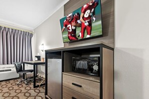 A hotel room with a flat-screen TV mounted above a wooden cabinet, a microwave, a refrigerator, a desk with a chair, and a patterned carpet.