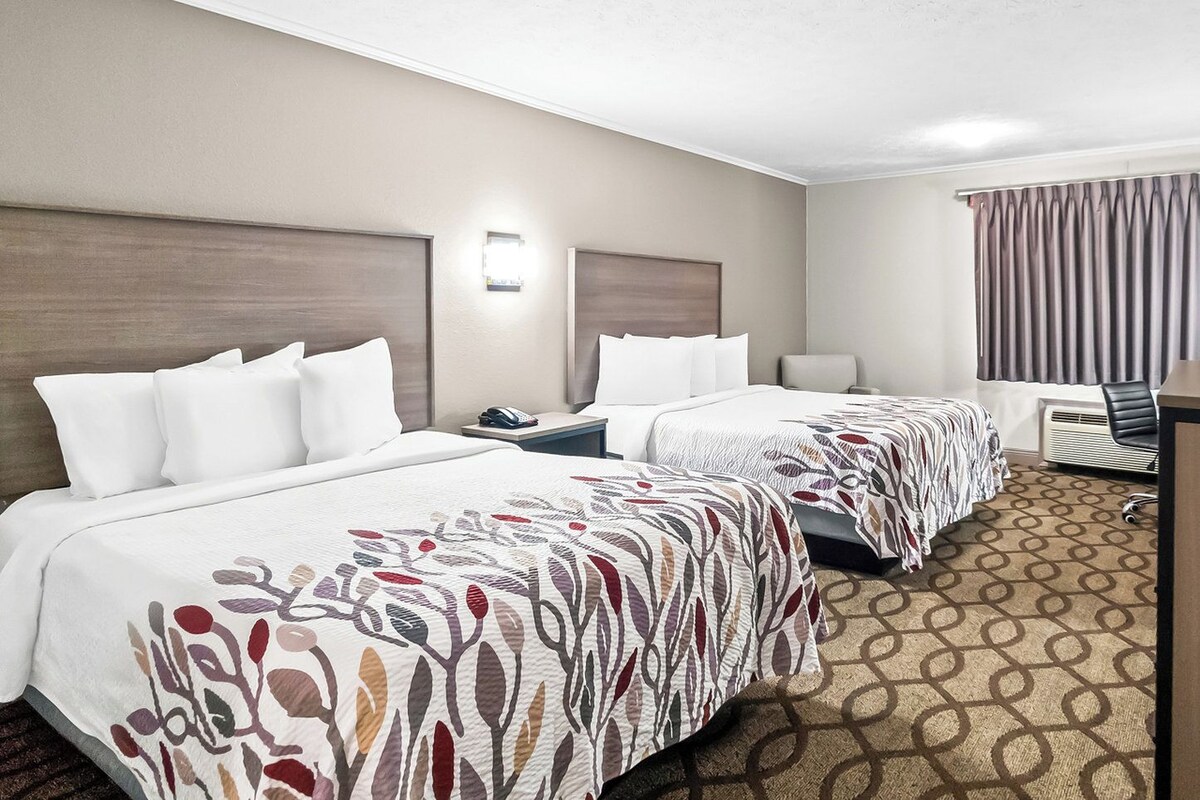 A hotel room with two beds, a desk, a chair, and a large window with curtains.