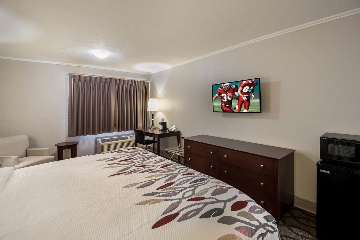 A hotel room with a large bed, a desk with a lamp, a TV on the wall, and a dresser.