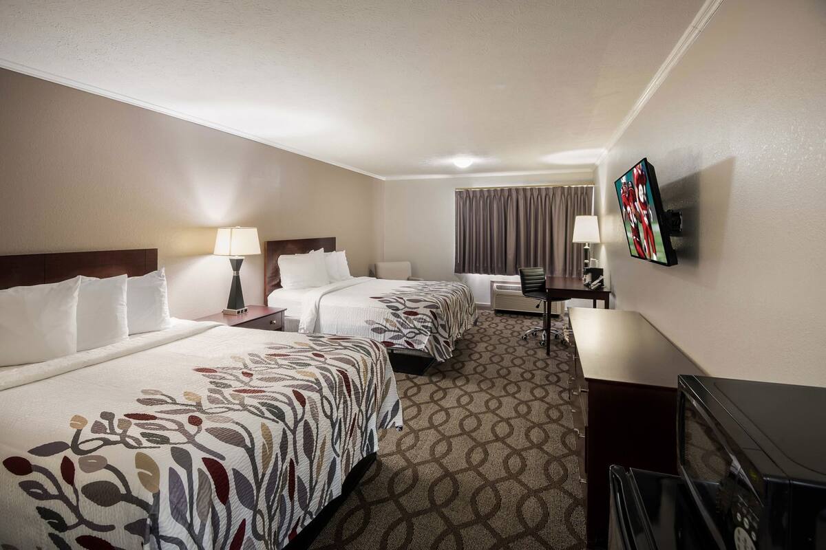 Une chambre d’hôtel avec deux lits, un bureau, une chaise, une télévision et une fenêtre avec des rideaux.