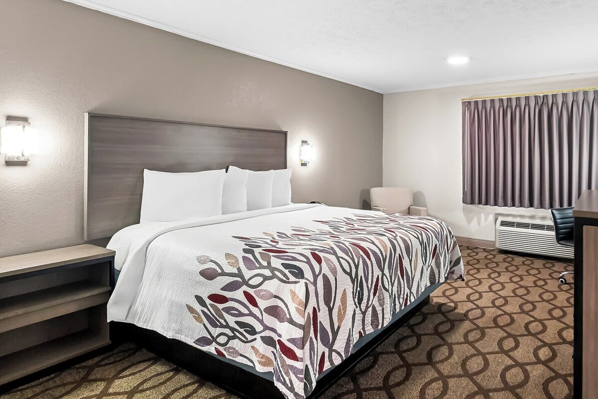 A hotel room with a bed, bedside table, lamp, chair, and air conditioning unit.