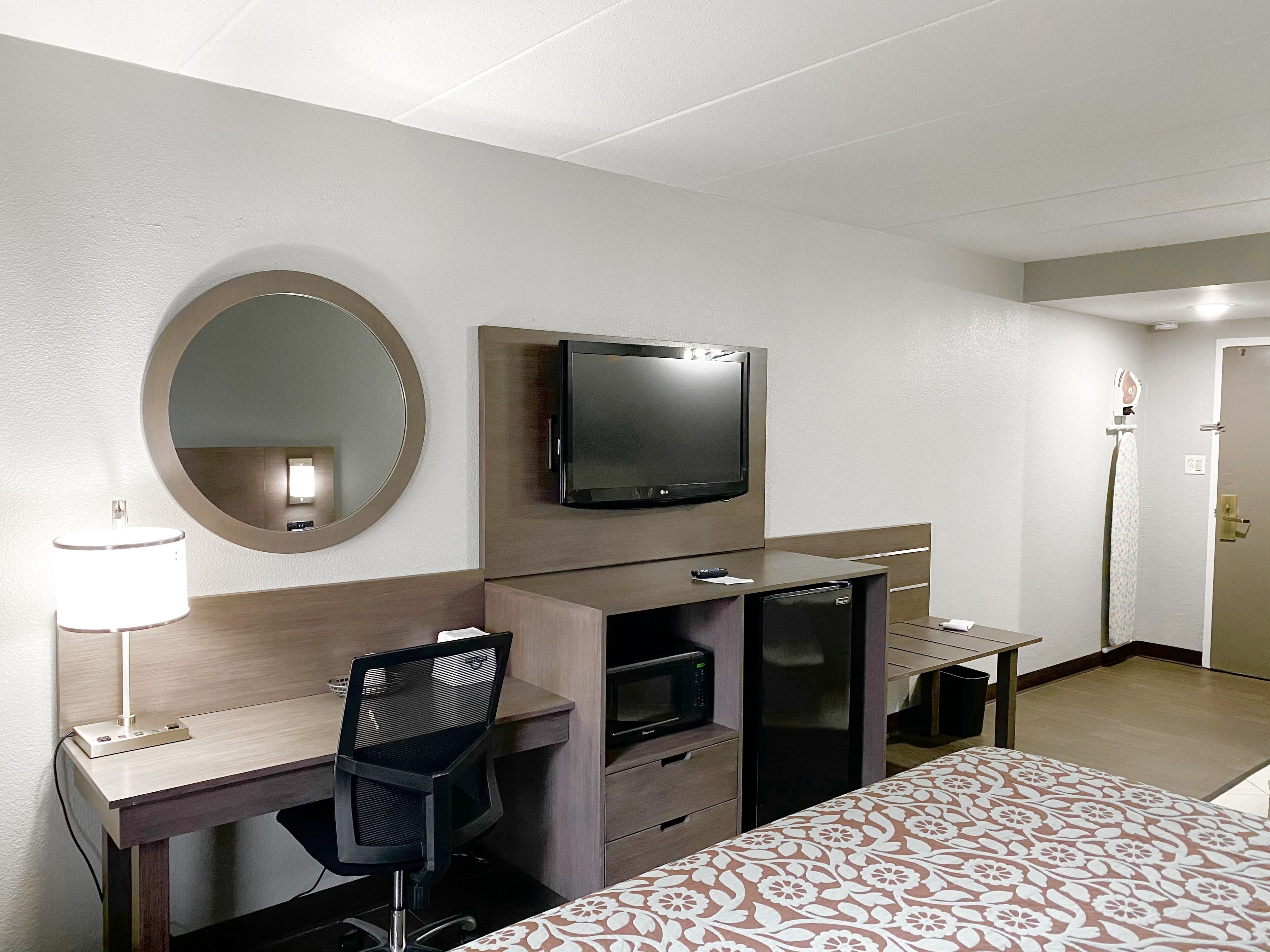 A hotel room with a bed, a desk with a chair, a television, a mirror, a microwave, and a refrigerator.