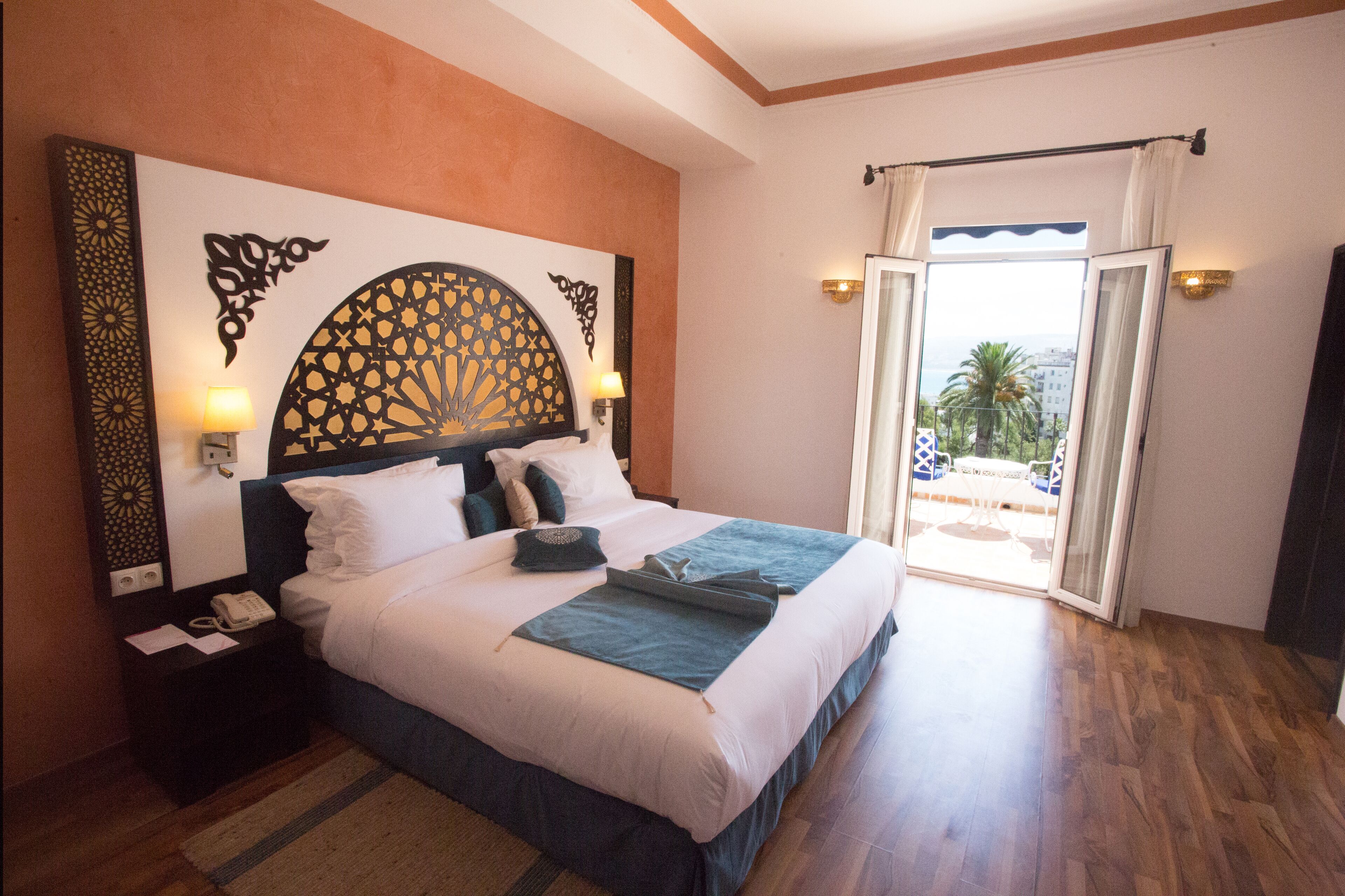 A hotel room with a large bed, a decorative headboard, bedside lamps, a telephone, and a view of the outdoors through a sliding door.