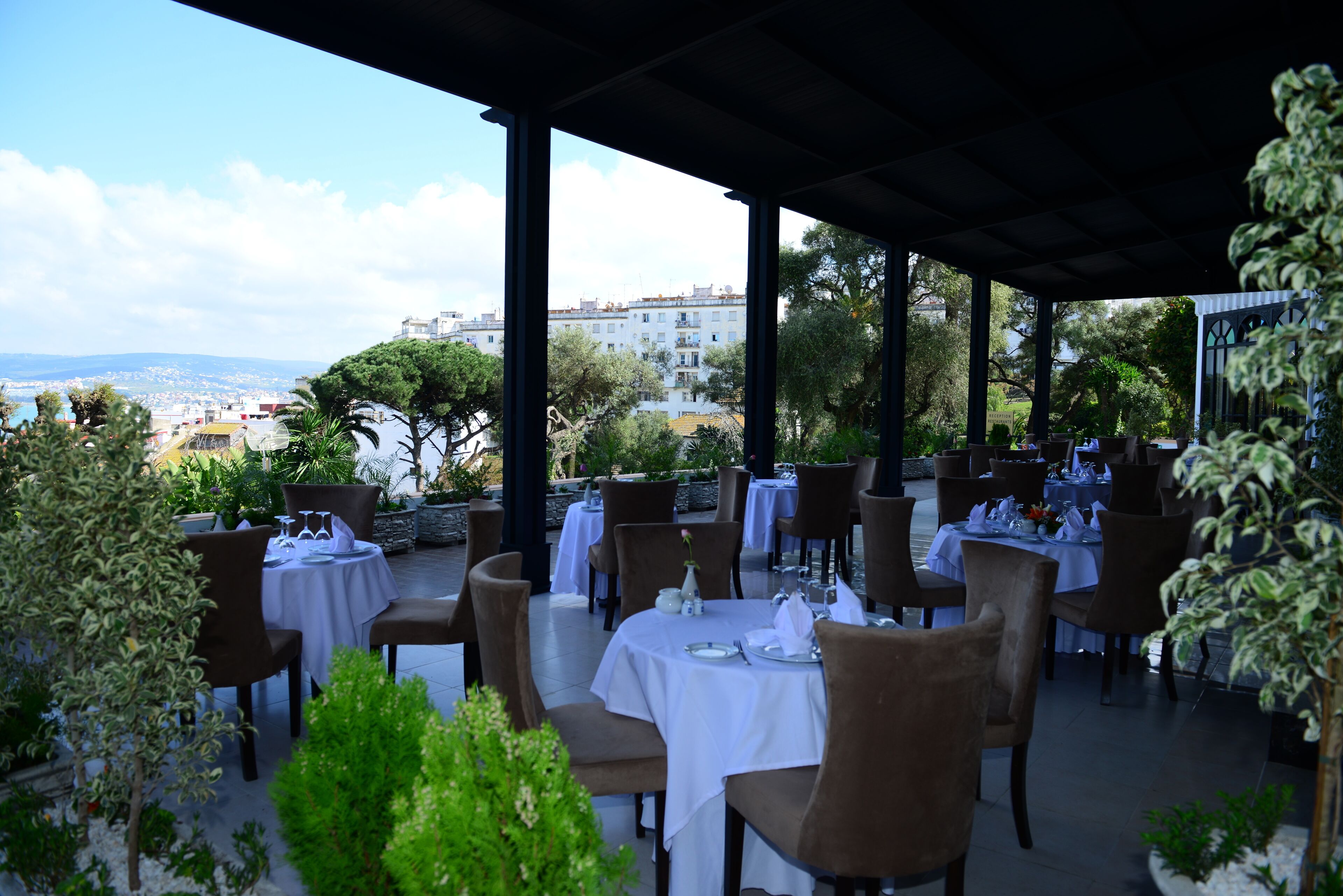 Un área de comedor al aire libre con mesas dispuestas para dos, rodeada de vegetación y con vistas a un paisaje urbano.