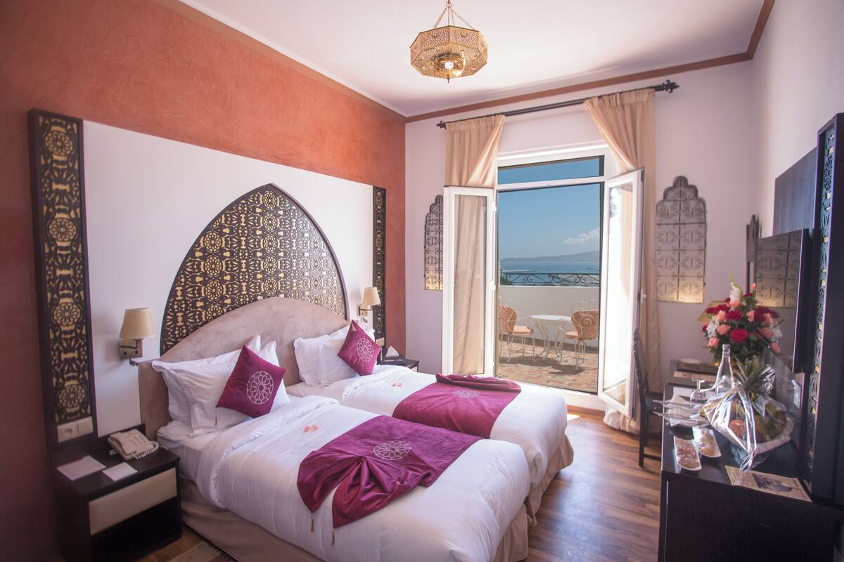 A hotel room with a large bed, a nightstand, a desk, a chair, a balcony with a view, and a decorative wall.