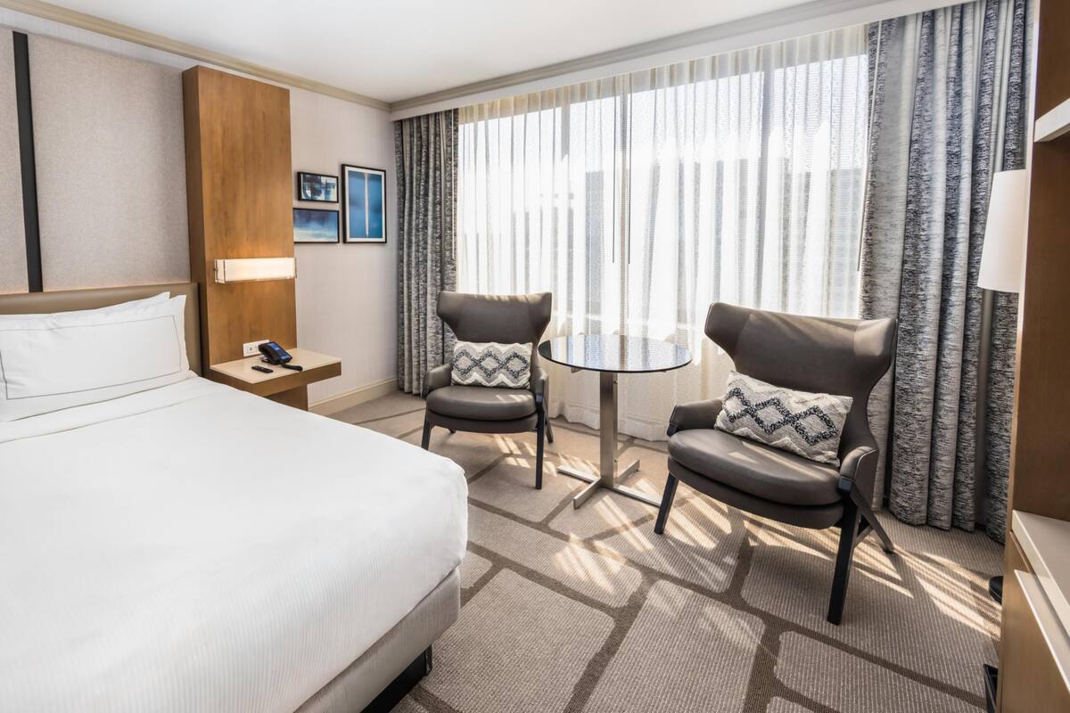 A hotel room with a large bed, two armchairs, a small table, a nightstand, and a window with curtains.