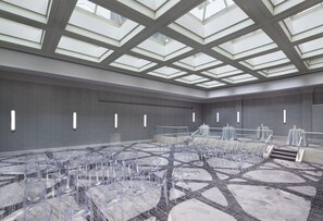 Una amplia sala de conferencias con techo alto y tragaluces, que cuenta con un escenario central con mesas y sillas dispuestas para un evento.