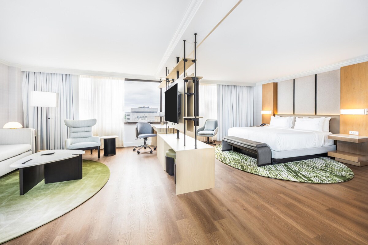 A hotel room with a bed, a desk, a chair, a sofa, a TV, and a window.