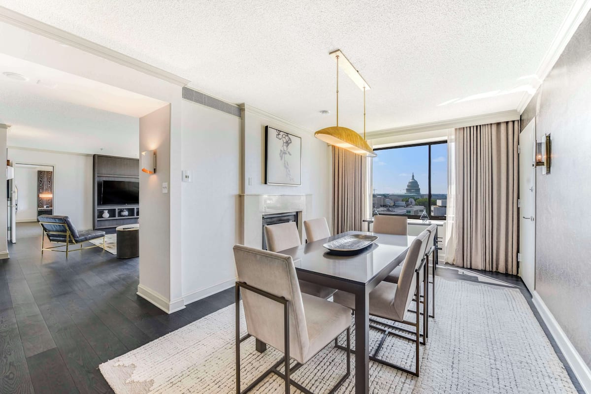 A modern hotel room with a dining area, a large window with a cityscape view, and a flat-screen TV.