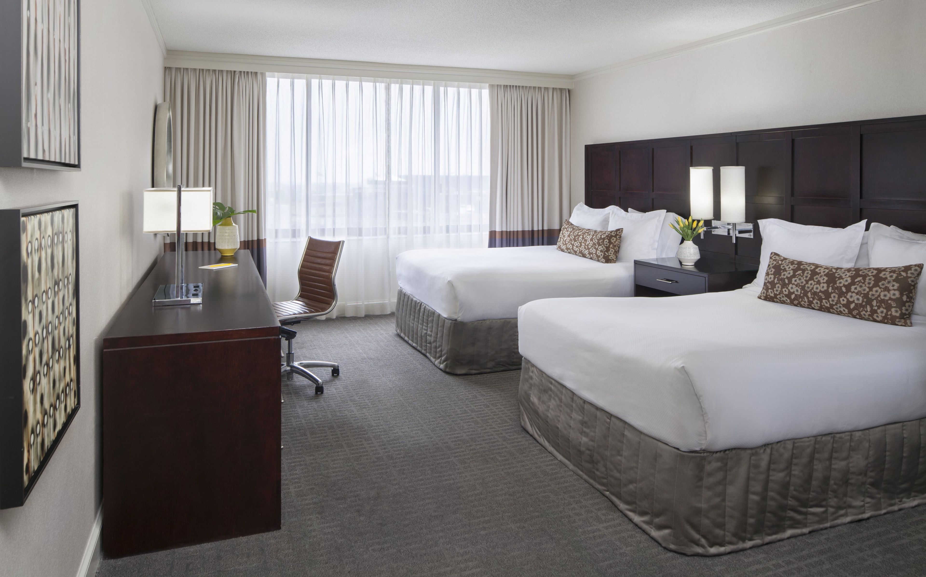 A hotel room with two beds, a desk, a chair, a nightstand, and a large window with curtains.