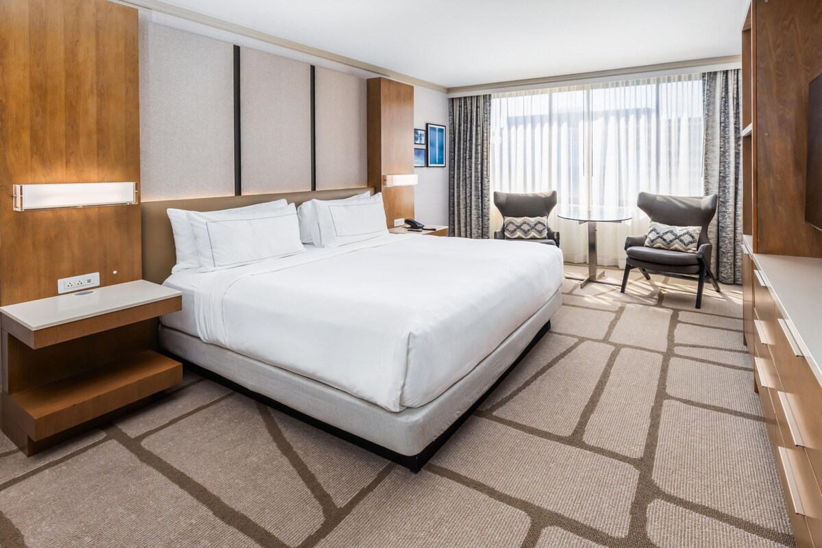A hotel room with a large bed, bedside tables, a desk, two chairs, and a window with curtains.
