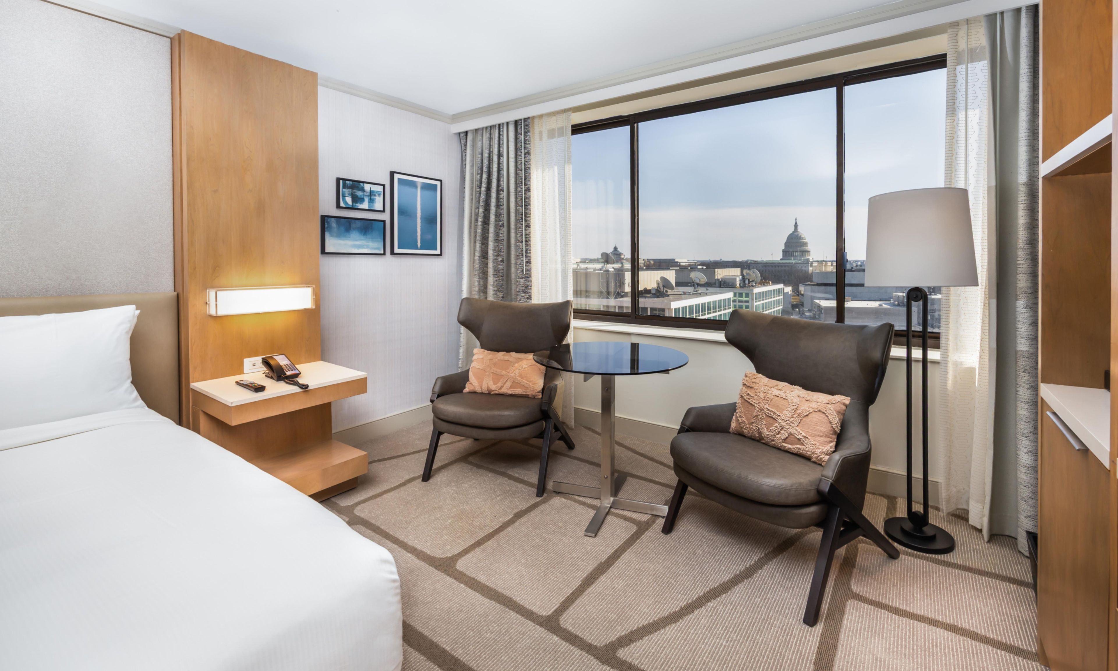 Habitación de hotel con cama, dos sillones, una mesita, un escritorio con teléfono y un amplio ventanal con vistas a la ciudad.