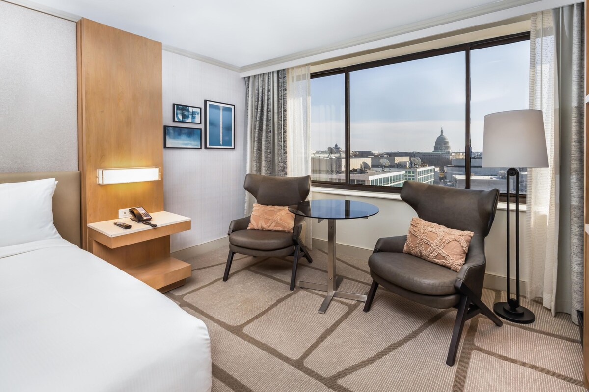 A hotel room with a bed, two armchairs, a small table, a desk with a phone, and a large window with a city view.