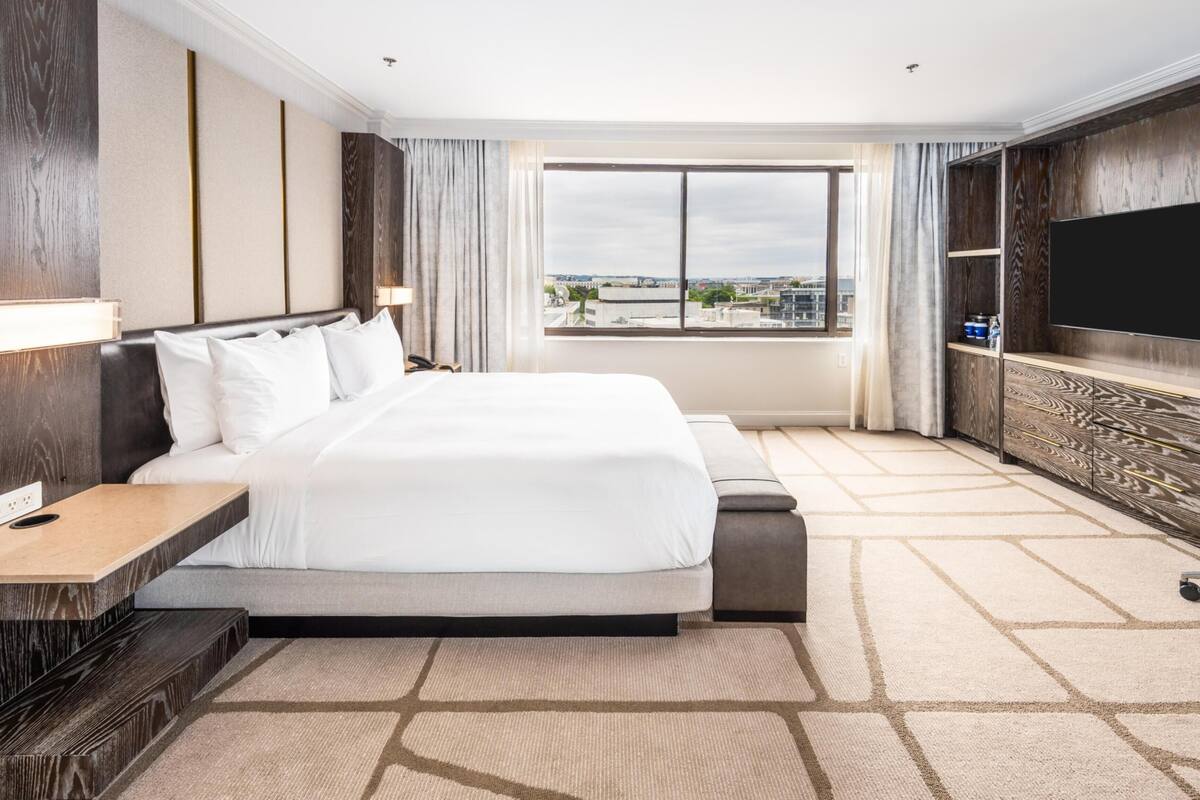 A hotel room with a large bed, a TV, a desk, and a view of the city.