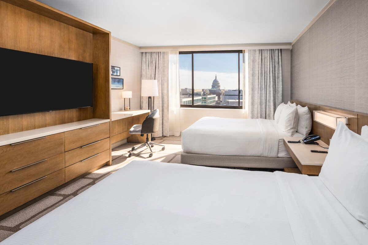 A hotel room with a large flat-screen TV, a desk with a lamp, two beds, a nightstand, and a view of a cityscape.