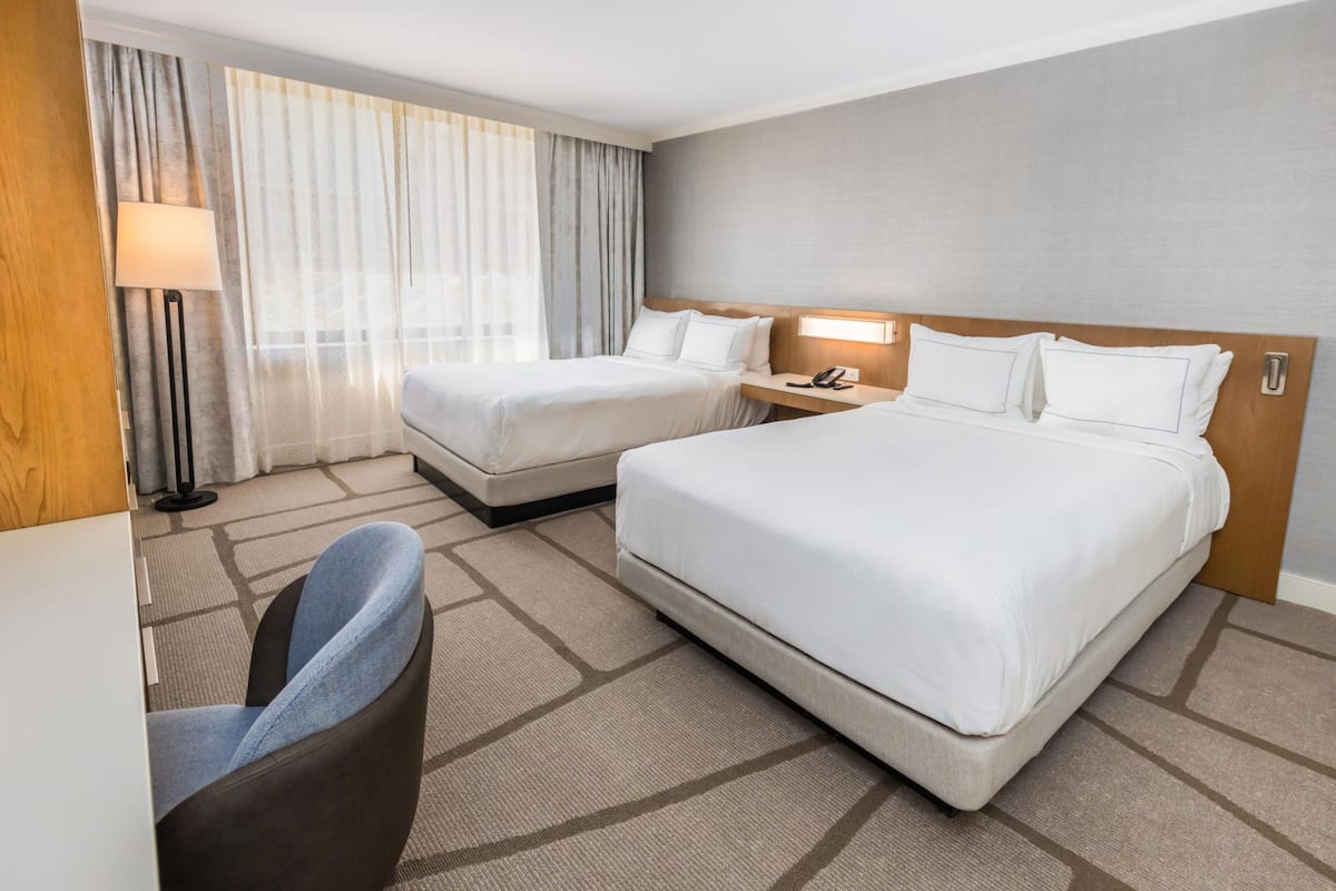 A hotel room with two beds, a desk, a chair, and a lamp.