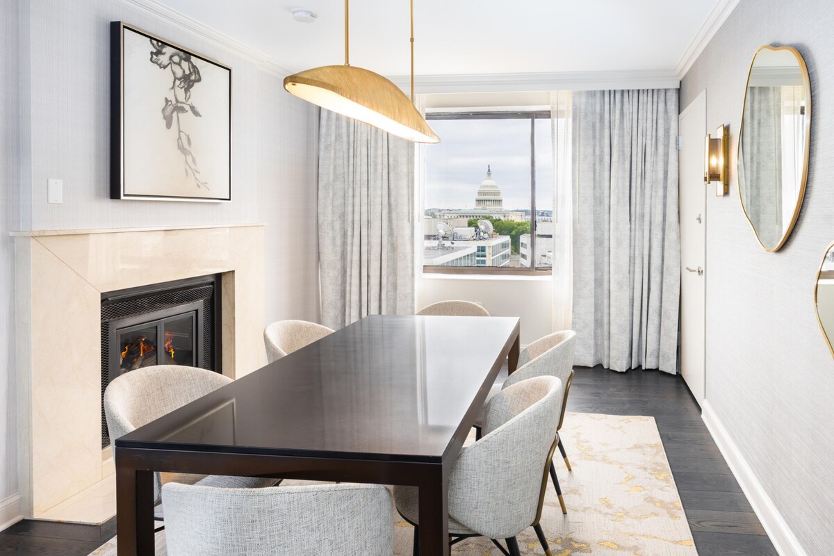 A modern dining room with a marble fireplace, a large dining table, and chairs. There are two large mirrors on the wall, a chandelier, and a view of the Capitol building through the window.