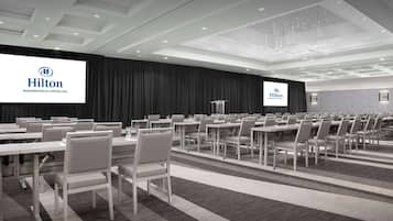 A conference room with multiple tables and chairs, a large screen displaying the Hilton logo, and a modern ceiling design.