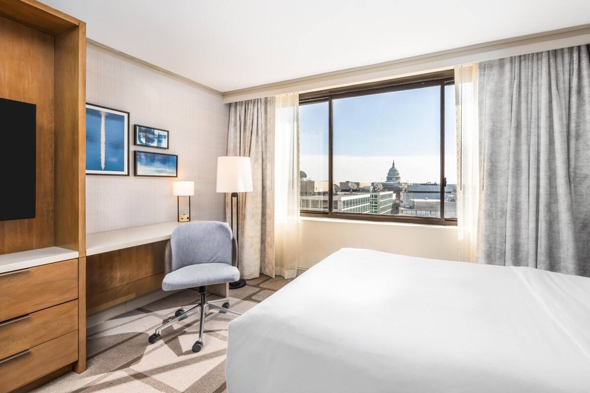 A hotel room with a bed, a desk, a chair, a TV, and a window with a cityscape view.