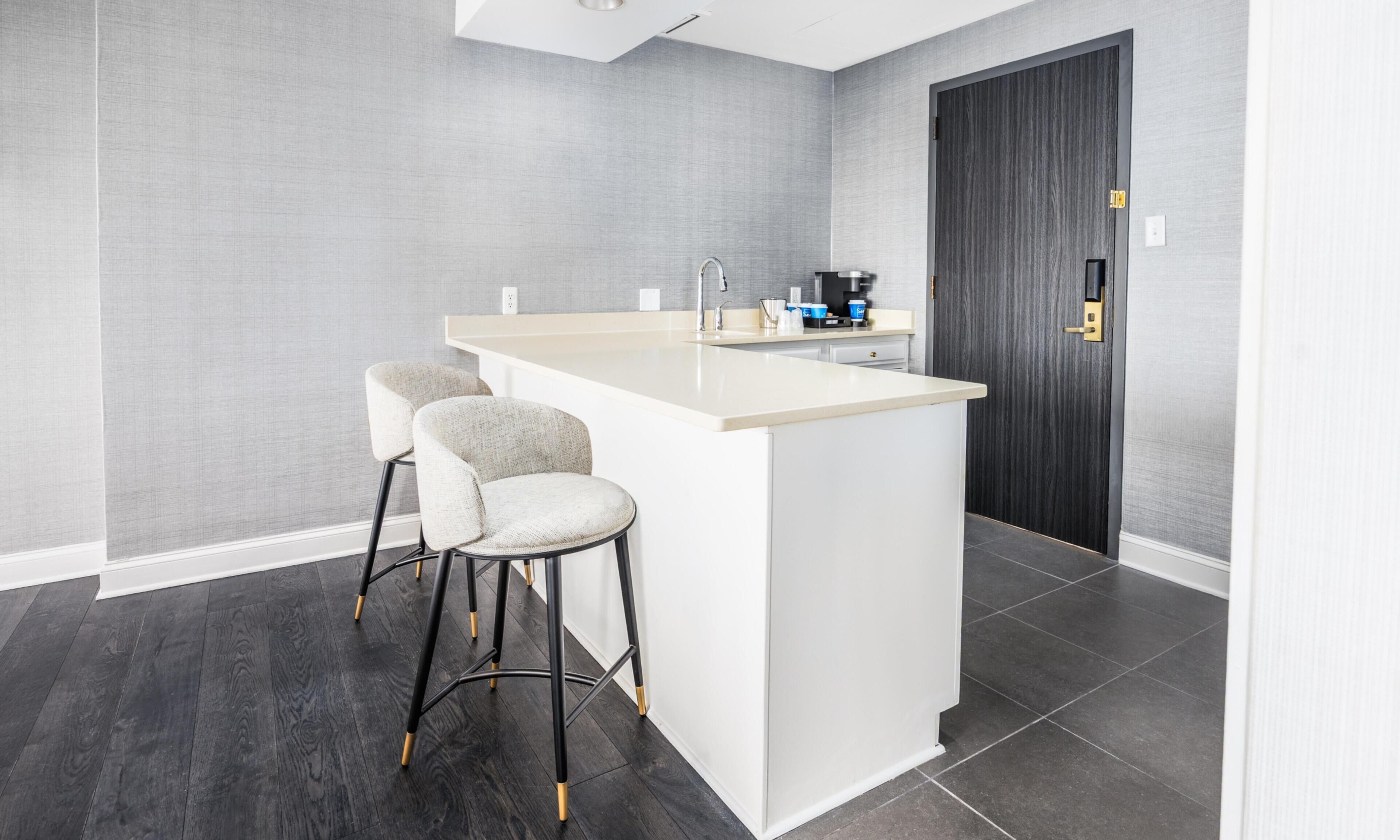 A modern kitchen with a white island, two bar stools, a dark wooden door, and grey textured walls.