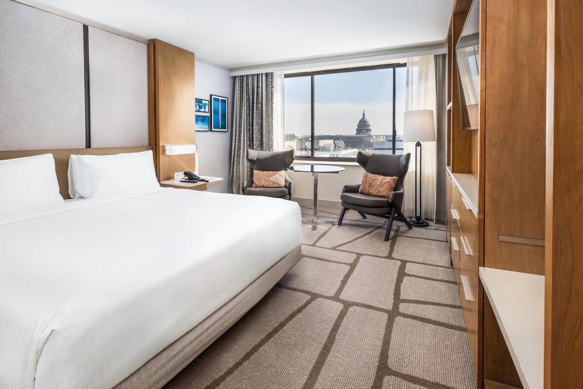 A hotel room with a large bed, two armchairs, a desk, and a view of a building through the window.