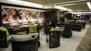 A modern hotel lobby with a geometric wall mural, multiple seating areas with sofas and chairs, and a bar area in the background.