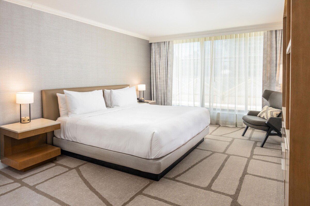A hotel room with a bed, bedside lamps, a chair, and a window with curtains.